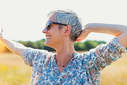 Eine Frau mit kurzem grauem Haar, die sich draussen in der Sonne streckt. Sie trägt eine Sonnenbrille und eine gemusterte Bluse.
