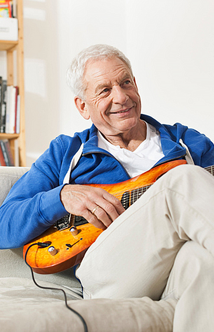 Ein lächelnder älterer Mann mit blauer Sweatjacke und beiger Hose, der auf einem beigen Sofa sitzt und Gitarre spielt.