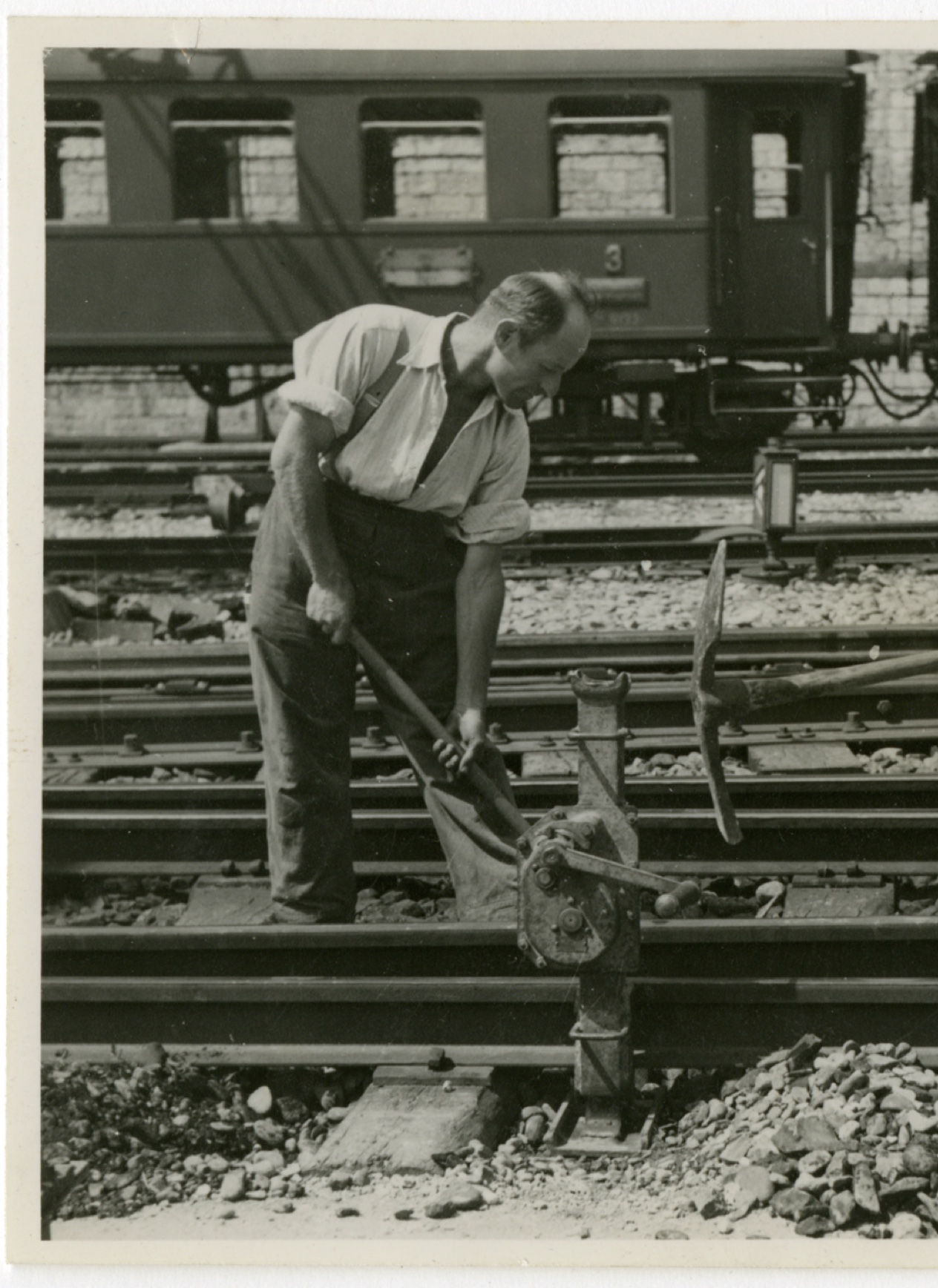 Alte schwarz-weiss Fotografie, auf der ein Handwerker abgebildet ist, der eine Zahnstangenwinde an einem Zuggleis bedient. Im Hintergrund befindet sich ein Eisenbahnwagen.